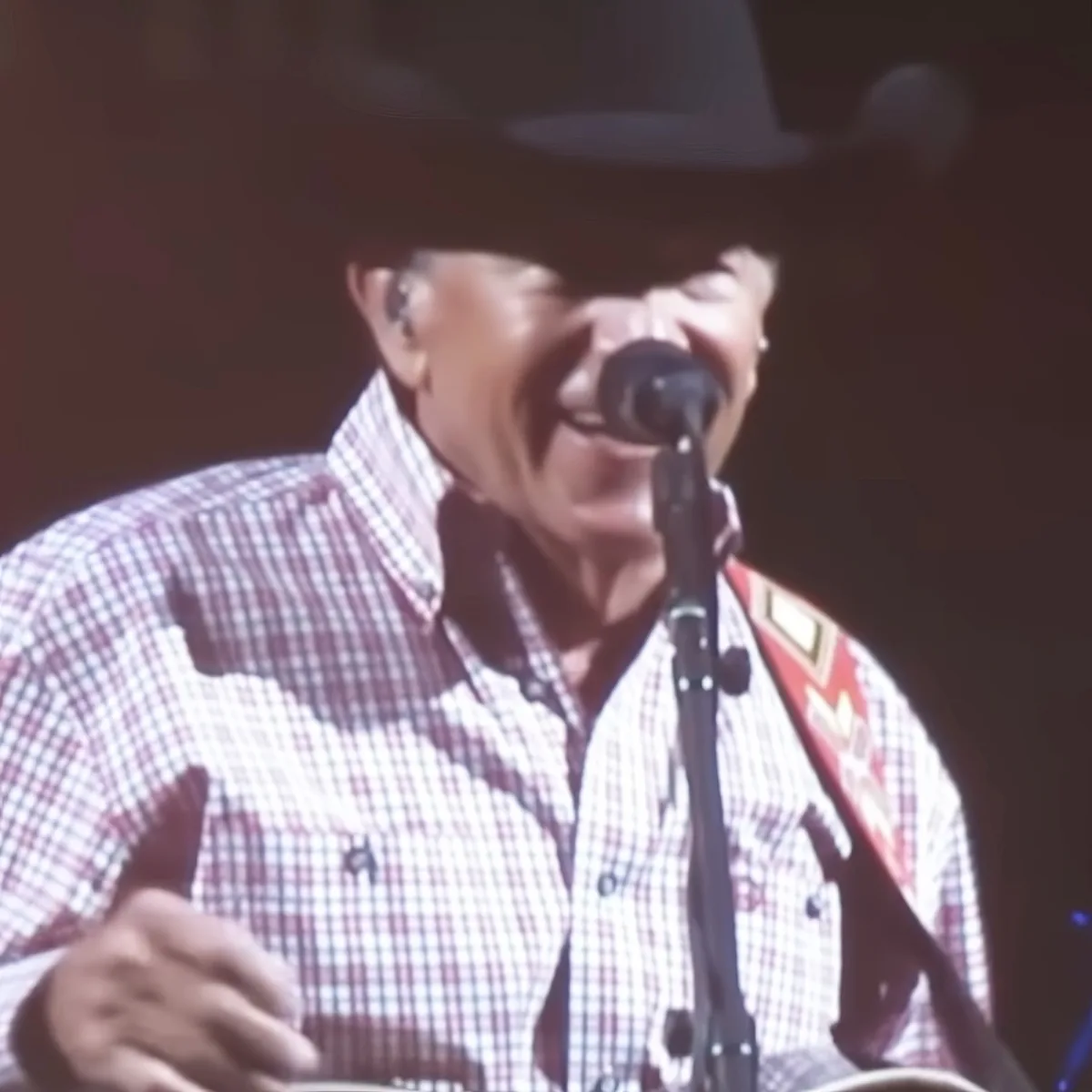 A man in a plaid shirt and cowboy hat sings into a microphone while playing a guitar.