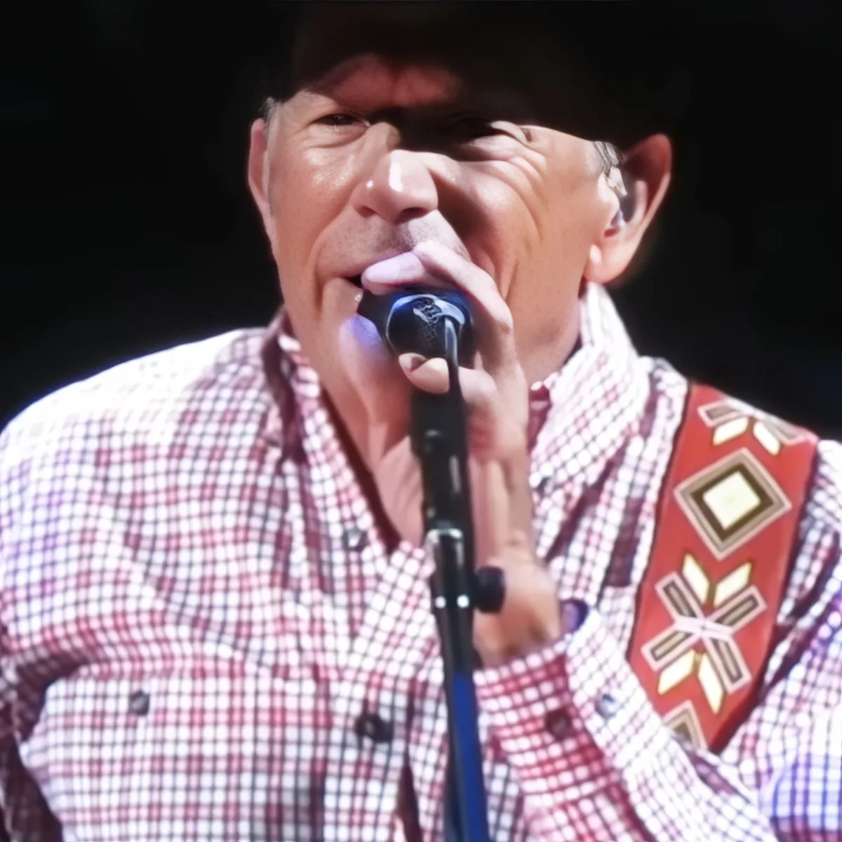 A man in a plaid shirt and cowboy hat sings into a microphone, wearing a decorative sash.