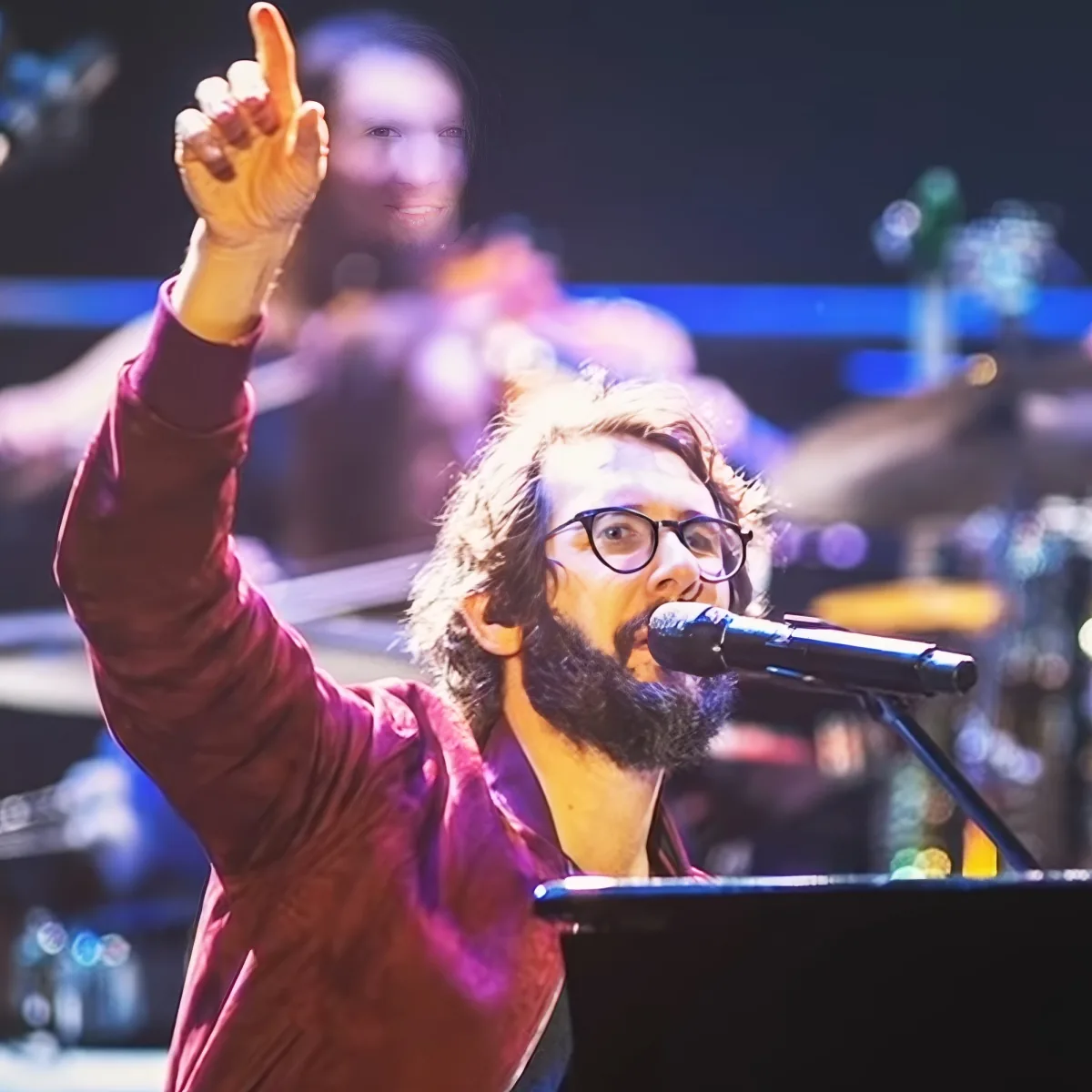 A man with a beard and glasses sings into a microphone, raising his hand in the air.