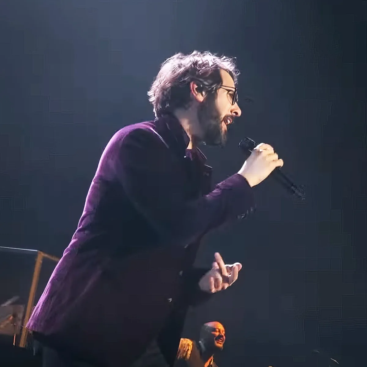 A man in a dark suit holds a microphone, singing passionately on stage.