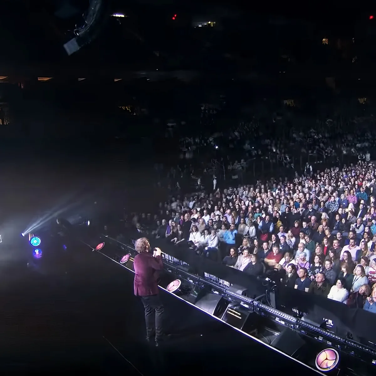 A performer stands on stage, illuminated by spotlights, addressing a large audience in a dimly lit venue.