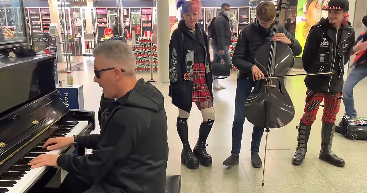 A man plays piano while a woman in a kilt and another in a black jacket with red plaid pants stand nearby, one holding a...