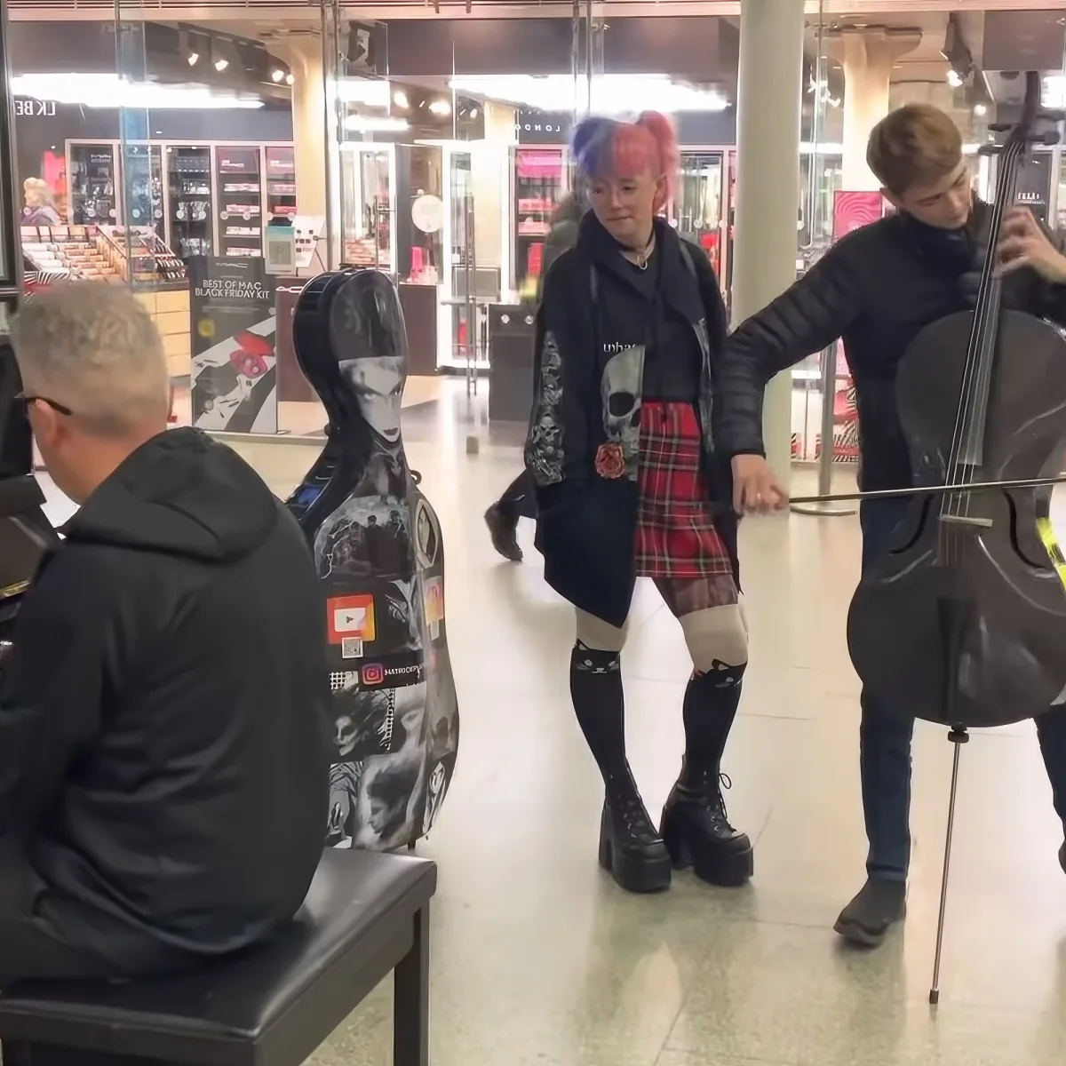 A person with colorful hair and a tartan skirt dances while a cellist plays in a shopping mall.