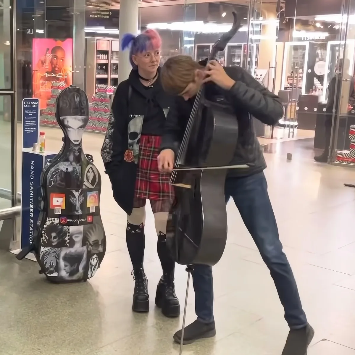 A person plays the cello in a public space, while another stands nearby, wearing a plaid skirt and knee-high socks.