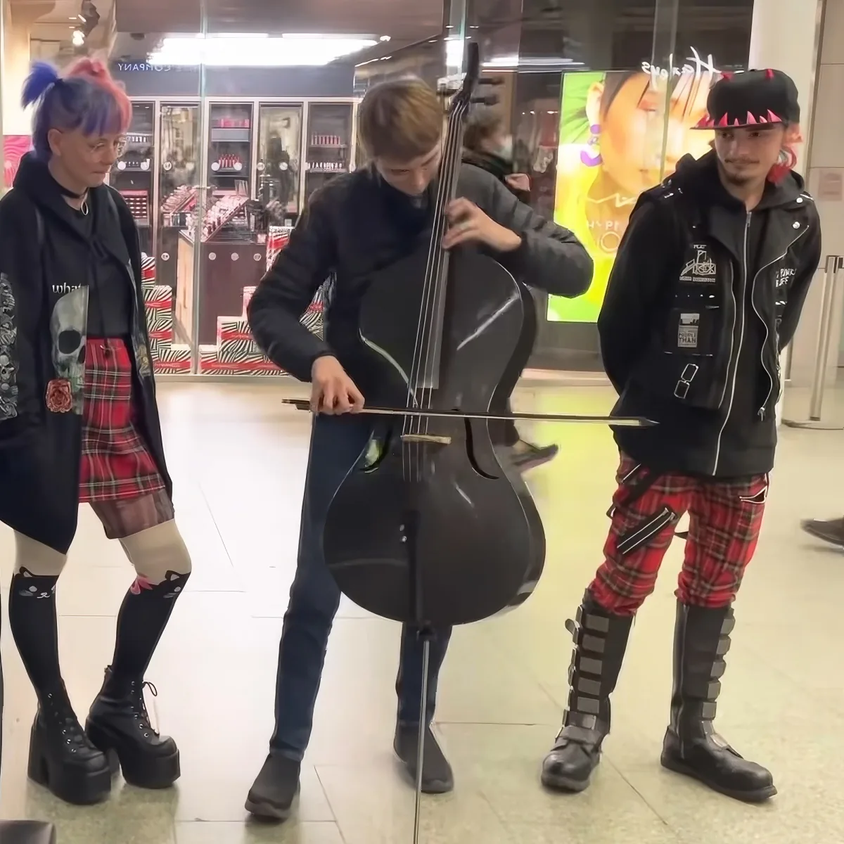 A person plays the cello while two others in punk-style attire watch in a shopping mall.