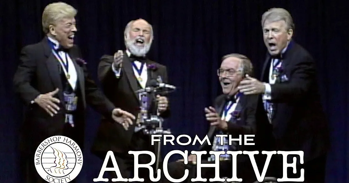 Four men in formal attire, one seated, all wearing medals, appear to be speaking or singing on stage.