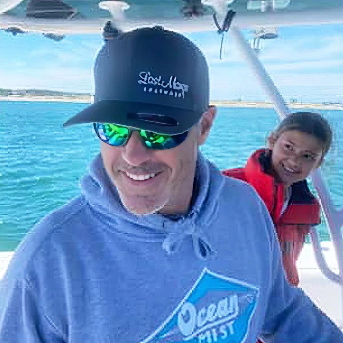 A man wearing a blue hoodie and sunglasses smiles on a boat with a woman in a life jacket behind him.