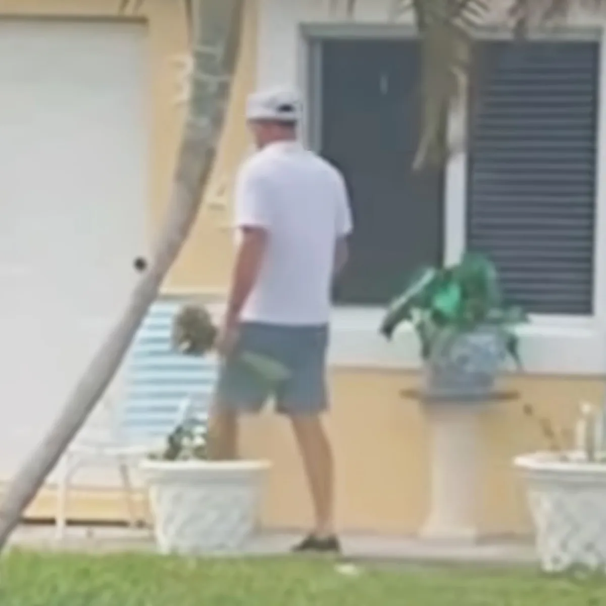 A man in a white shirt and hat walks past a house with a pool and plants.