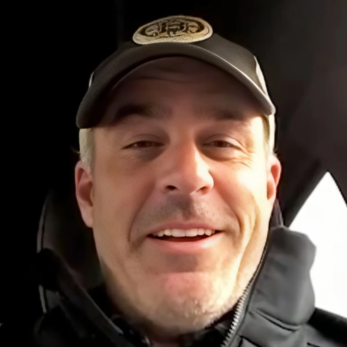 A smiling man wearing a cap and a jacket, with a dark background.