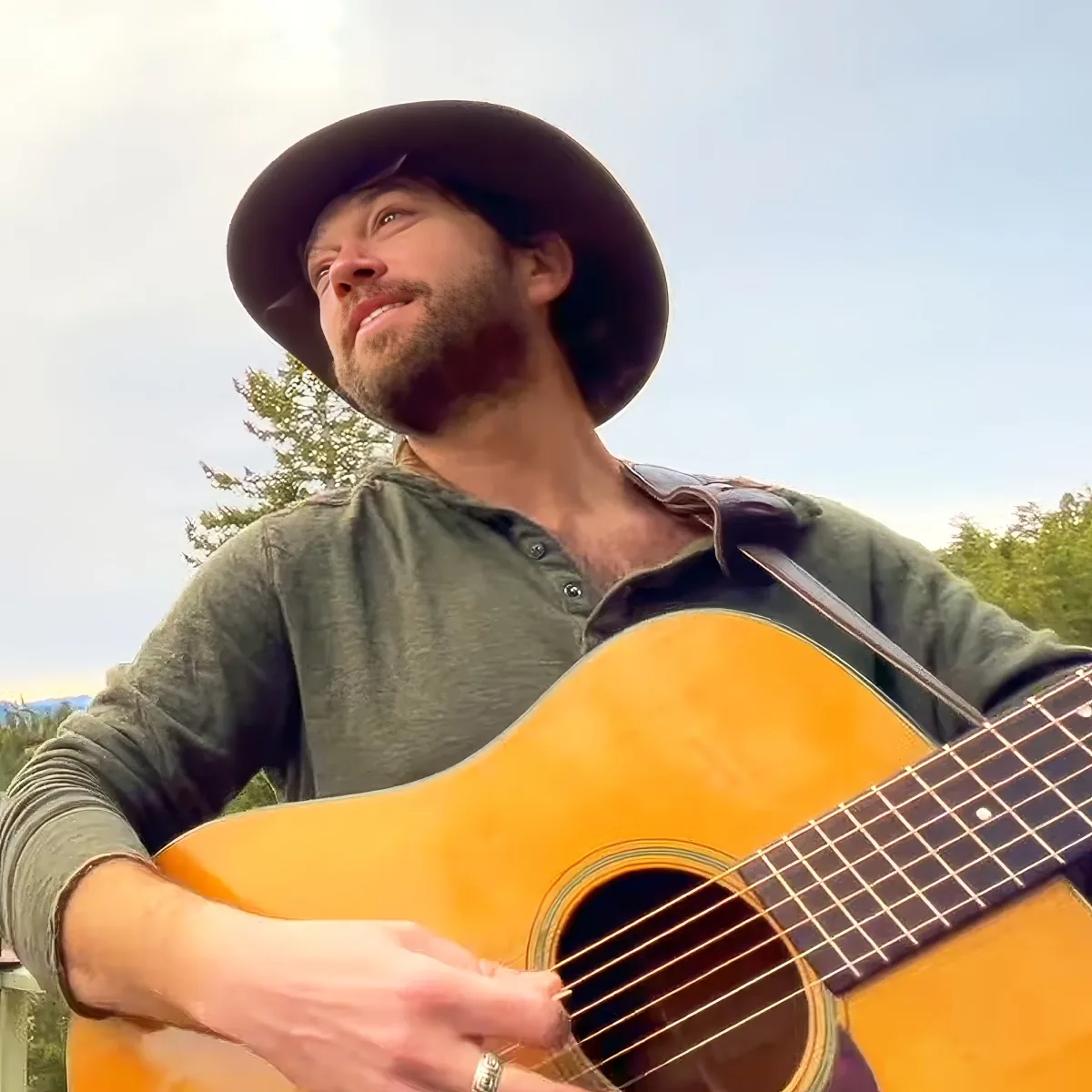 A man in a cowboy hat plays an acoustic guitar outdoors, surrounded by trees.