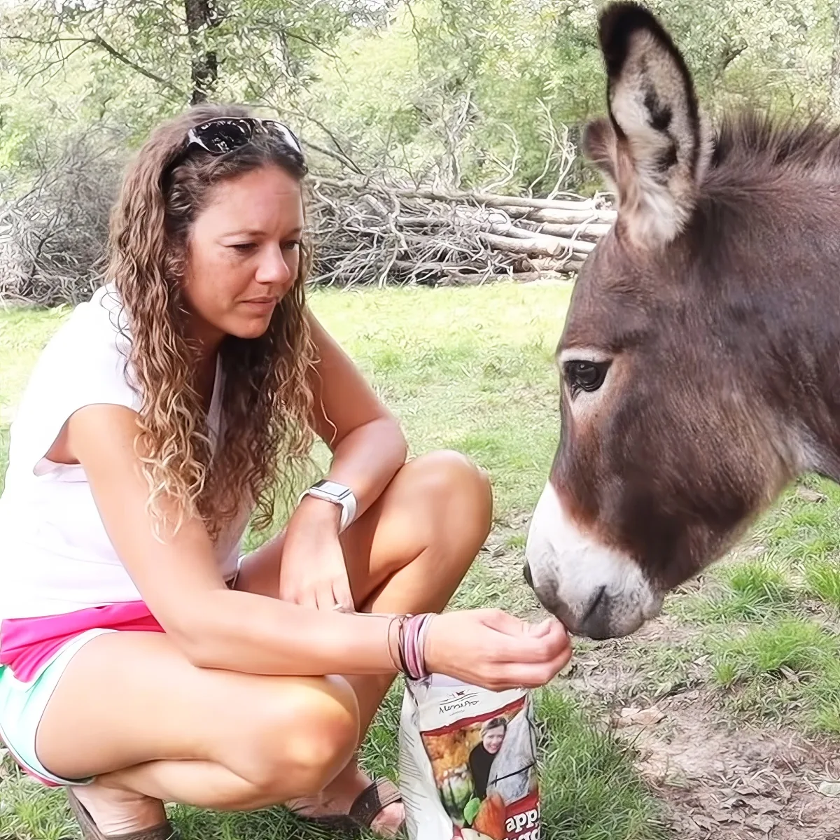 A woman crouches to feed a donkey, both in a grassy outdoor setting.