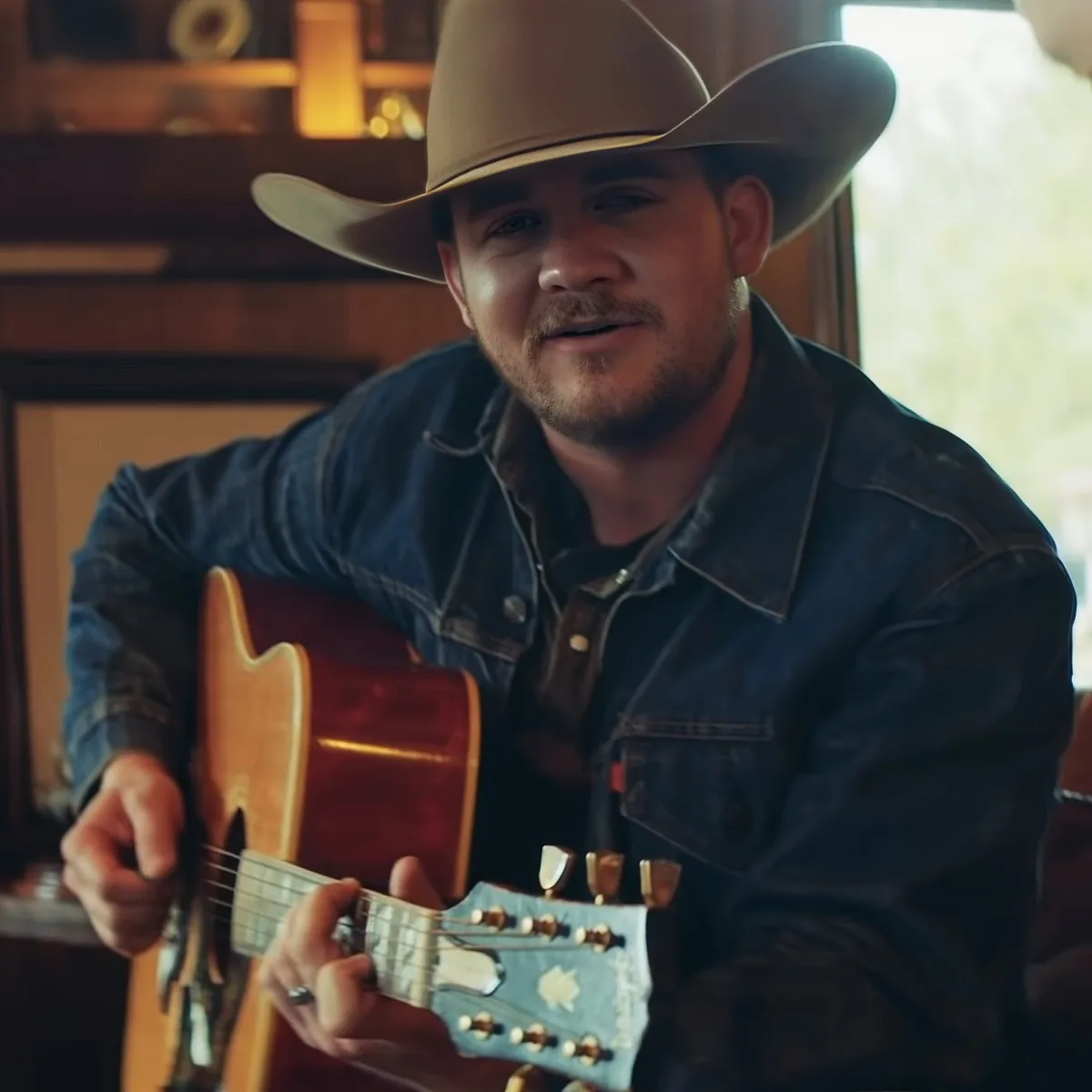 A man in a cowboy hat and denim jacket plays an acoustic guitar indoors.