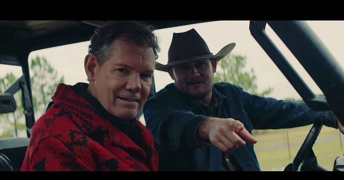 A man in a red patterned jacket sits in a vehicle, smiling, with another man in a cowboy hat behind him.