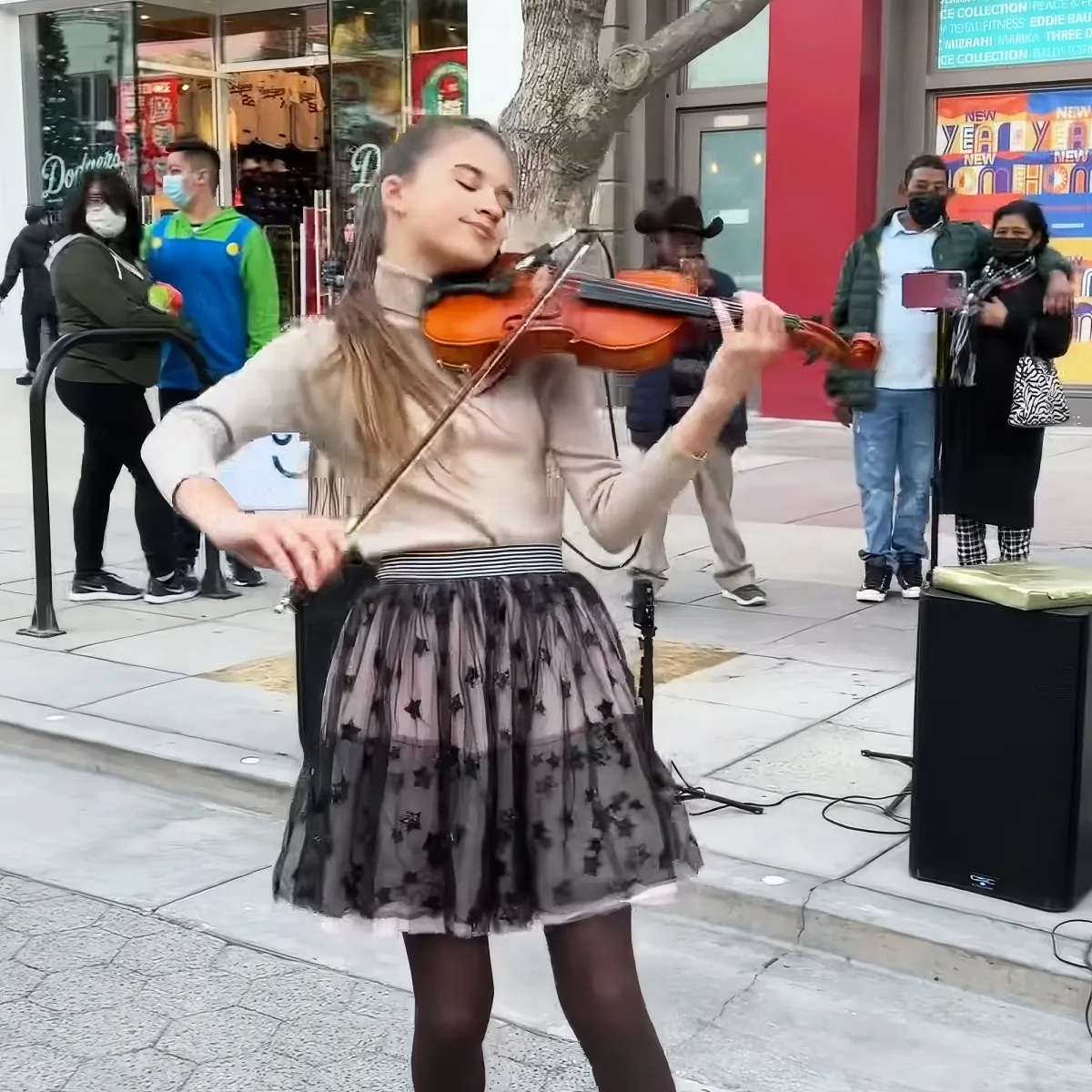 A girl in a tulle skirt plays the violin on a city sidewalk, surrounded by onlookers.
