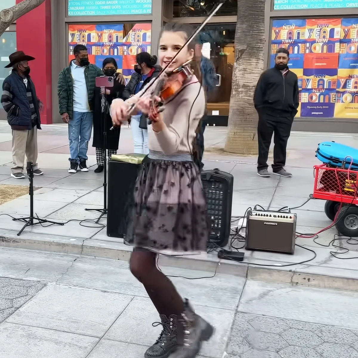 A young girl plays violin on a city sidewalk, surrounded by onlookers and musical equipment.