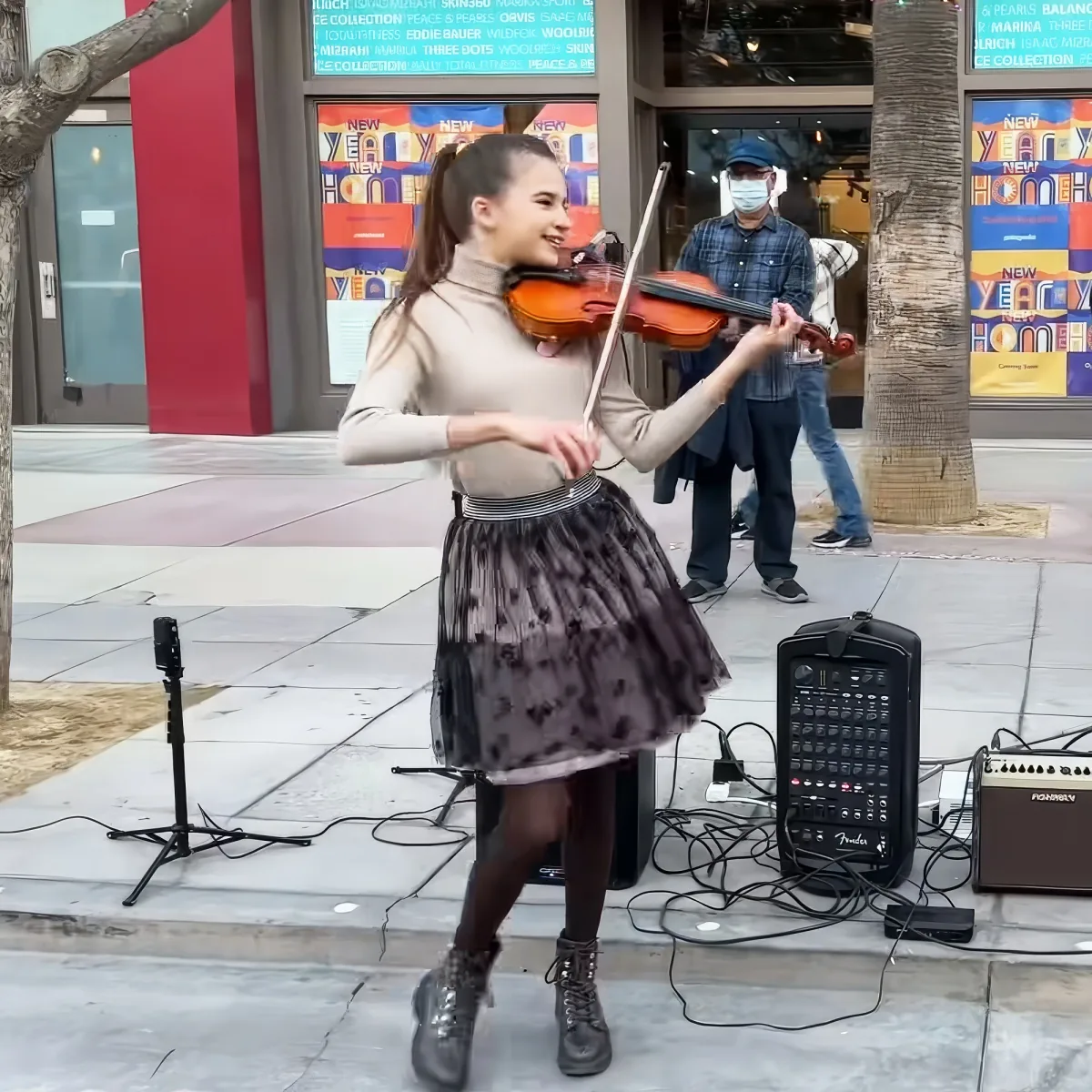 A young woman plays violin on a city sidewalk, surrounded by musical equipment and a passerby.
