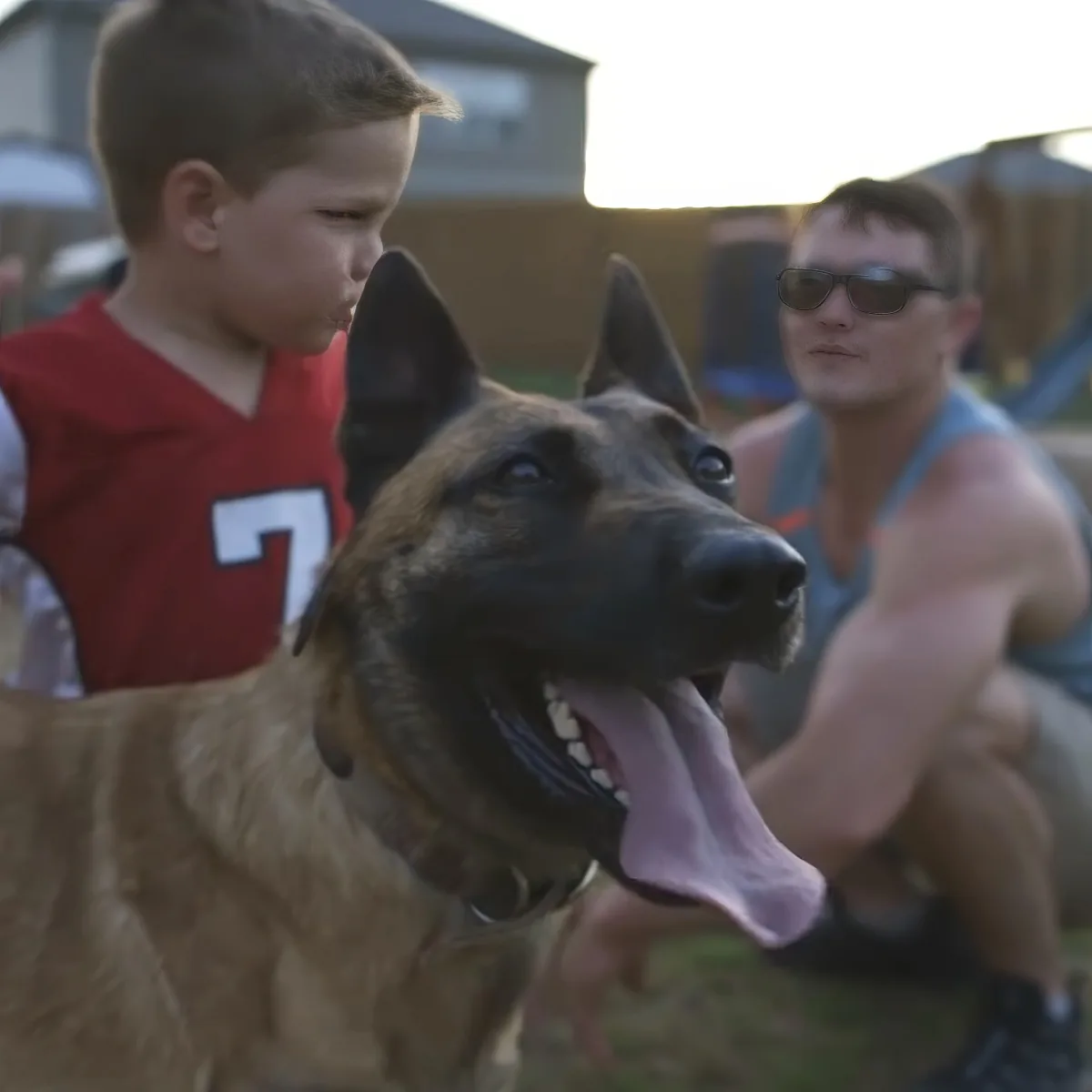 A German Shepherd with its tongue out, standing next to a boy in a red jersey and a man in sunglasses.