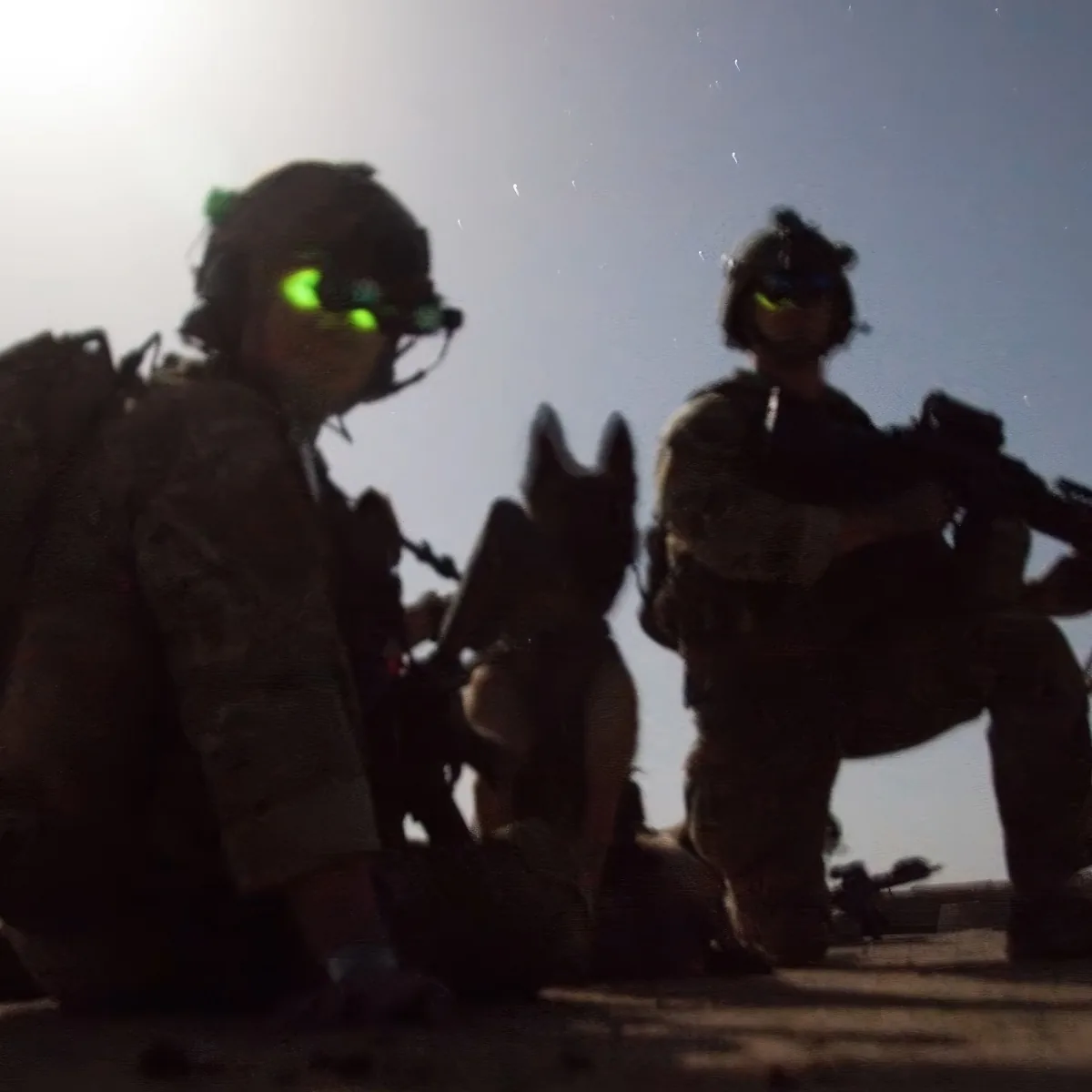 Two soldiers in night vision gear with a dog, silhouetted against a bright light.