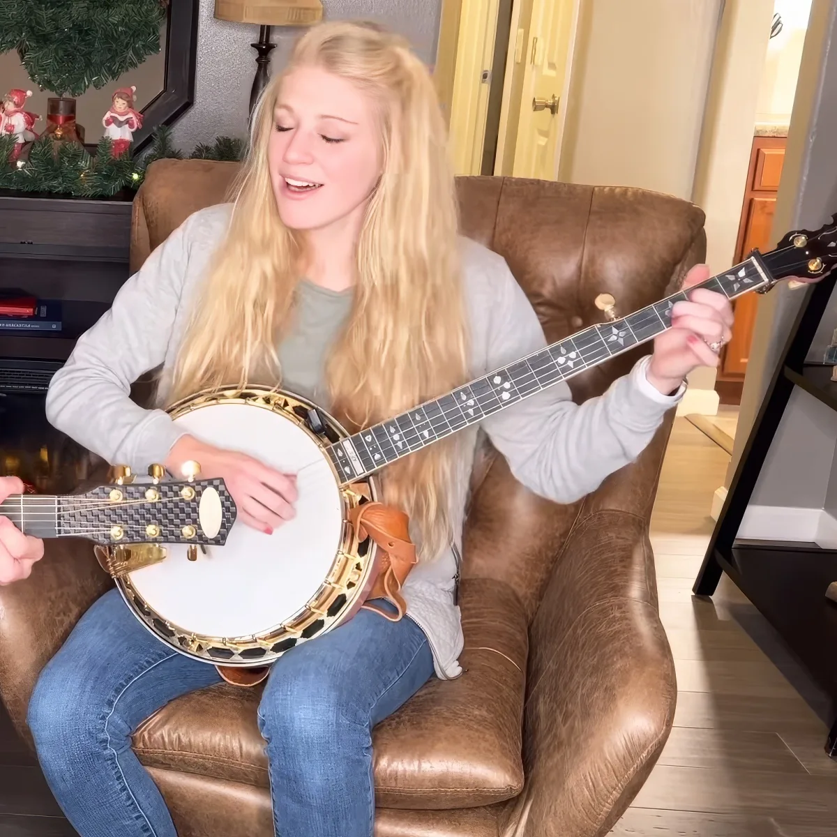 A person with long blonde hair plays a banjo while sitting on a brown leather couch.
