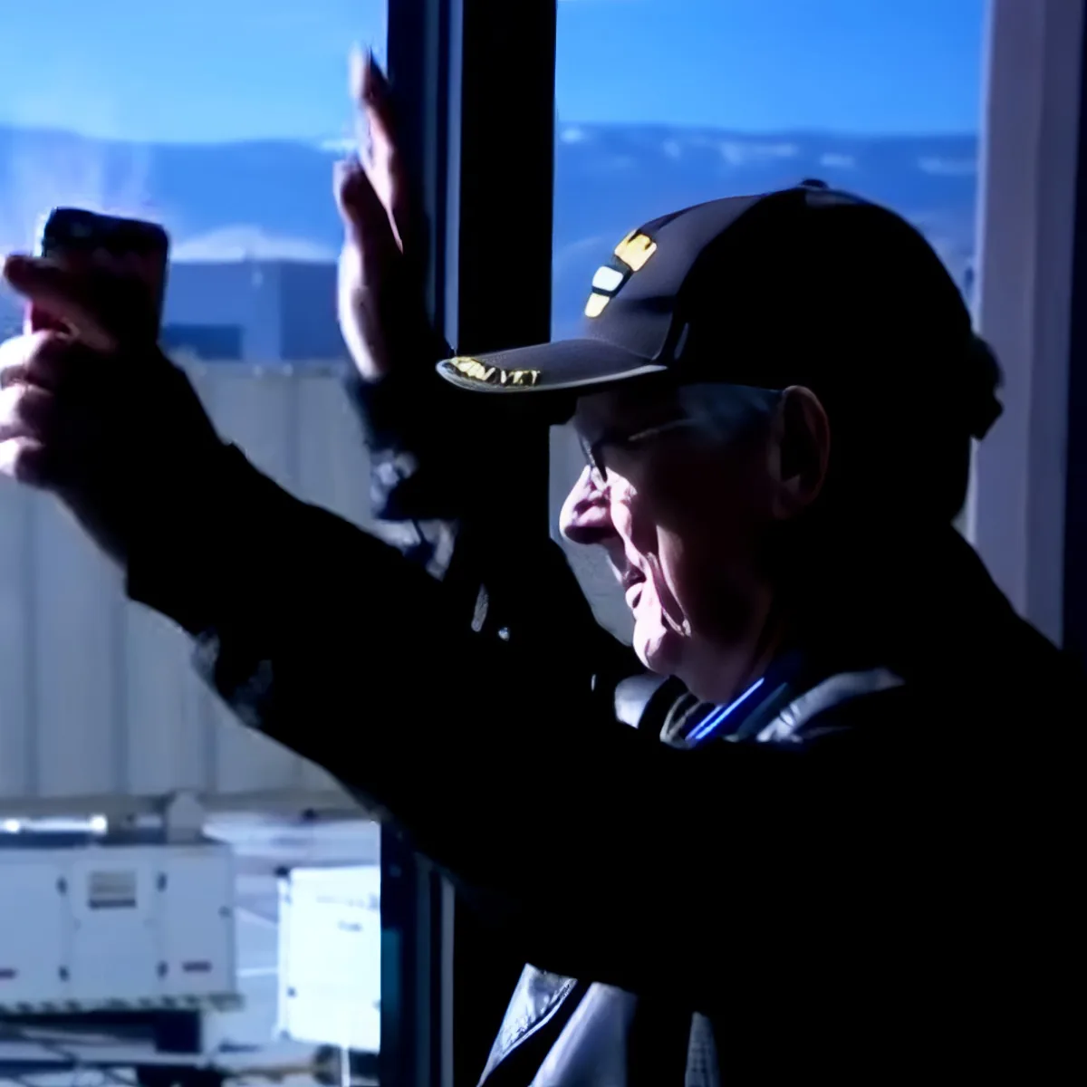 An elderly man wearing a cap and glasses is taking a photo with his phone by a window.