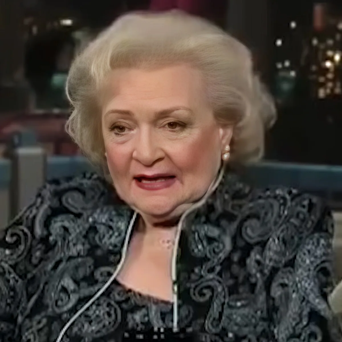 An elderly woman with white hair, wearing a black patterned jacket, sits in a chair, appearing to speak.