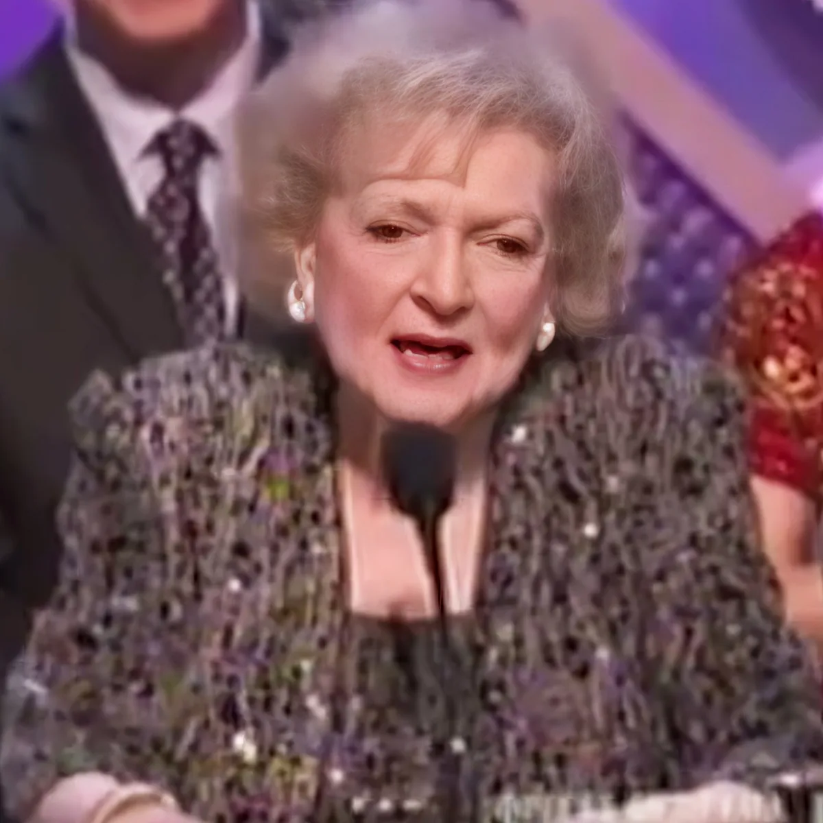 An older woman in a sequined dress speaks into a microphone at an awards ceremony.