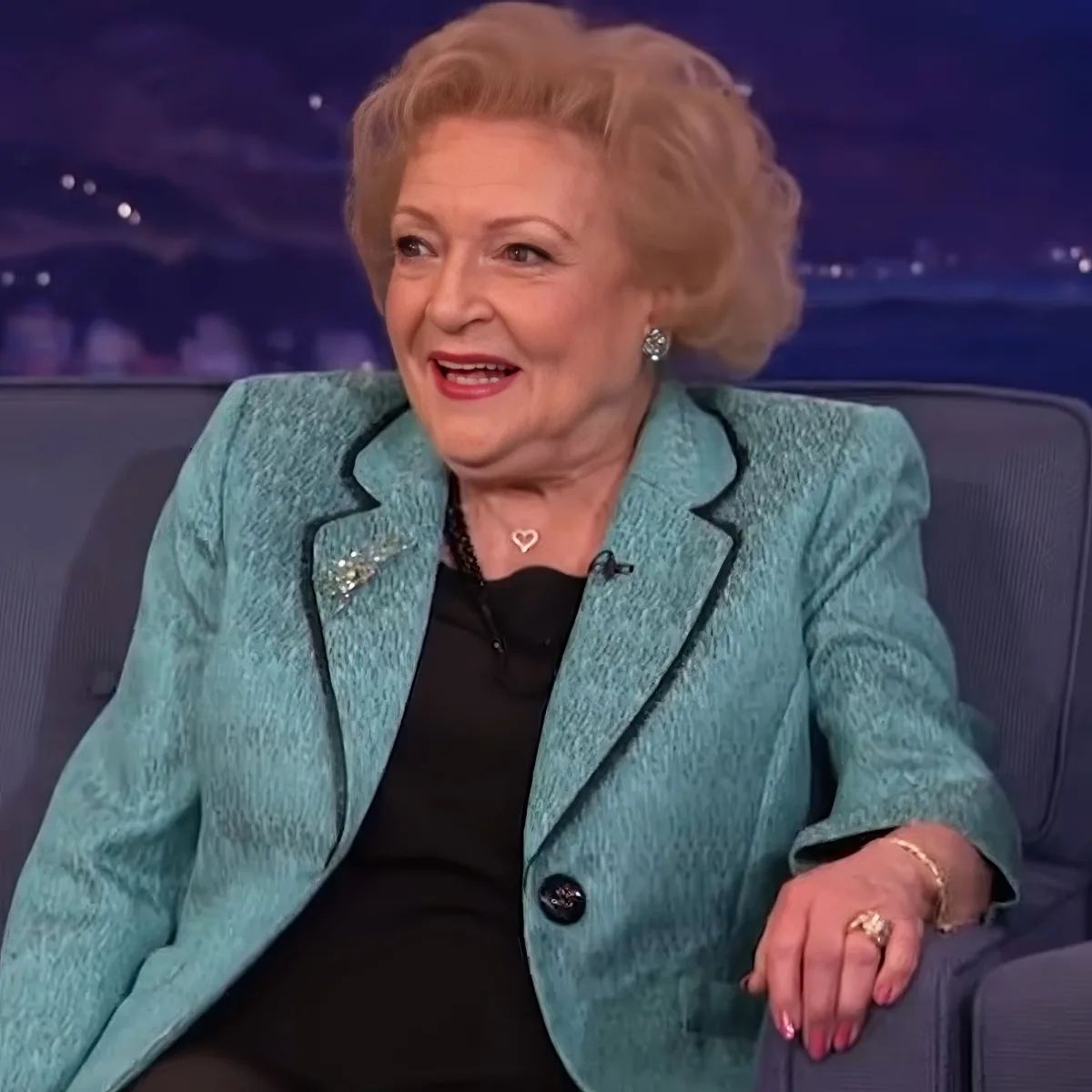 A woman in a teal blazer and black dress is seated, smiling, with a heart-shaped necklace and earrings.