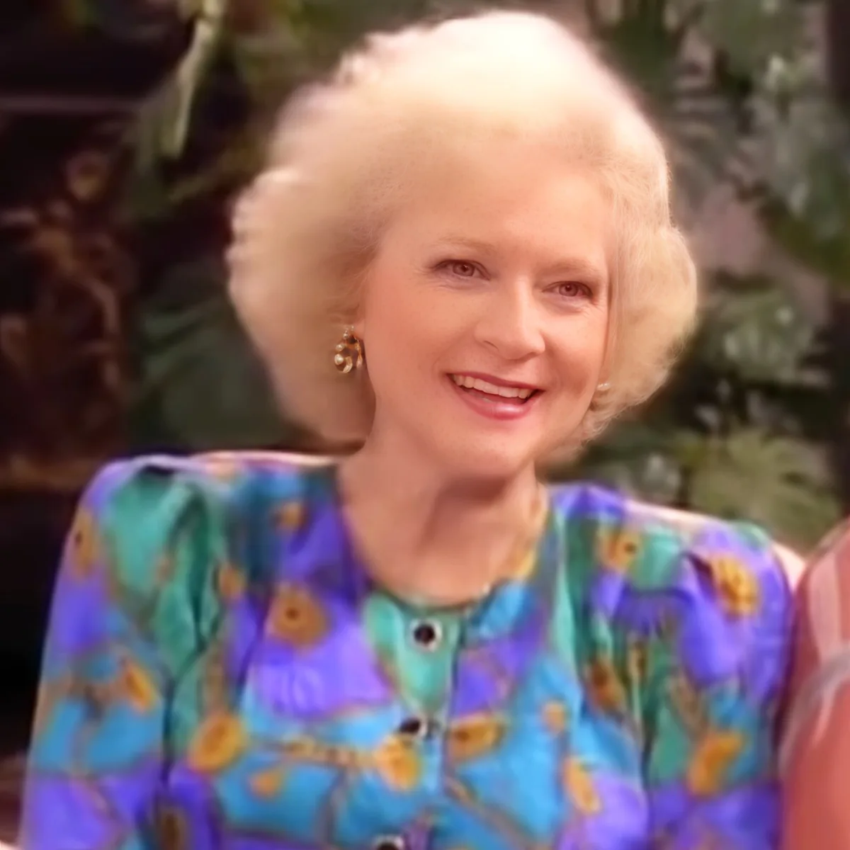 An elderly woman with white hair, wearing a colorful floral dress, smiles warmly.
