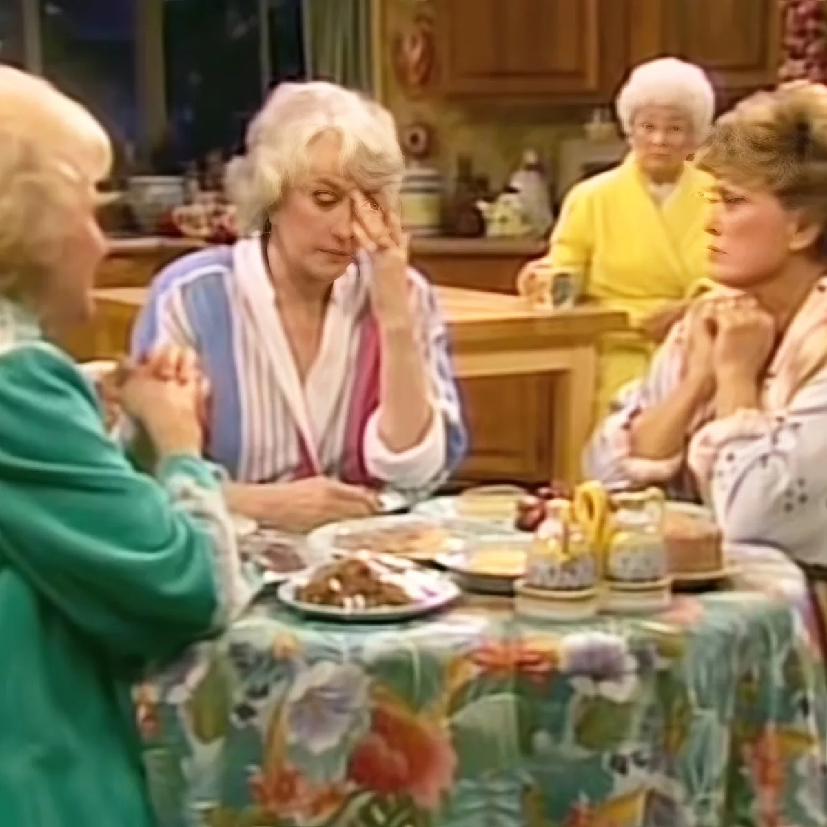 Four women in a kitchen setting, one in a green robe, another in a striped robe, and two others in yellow and white...