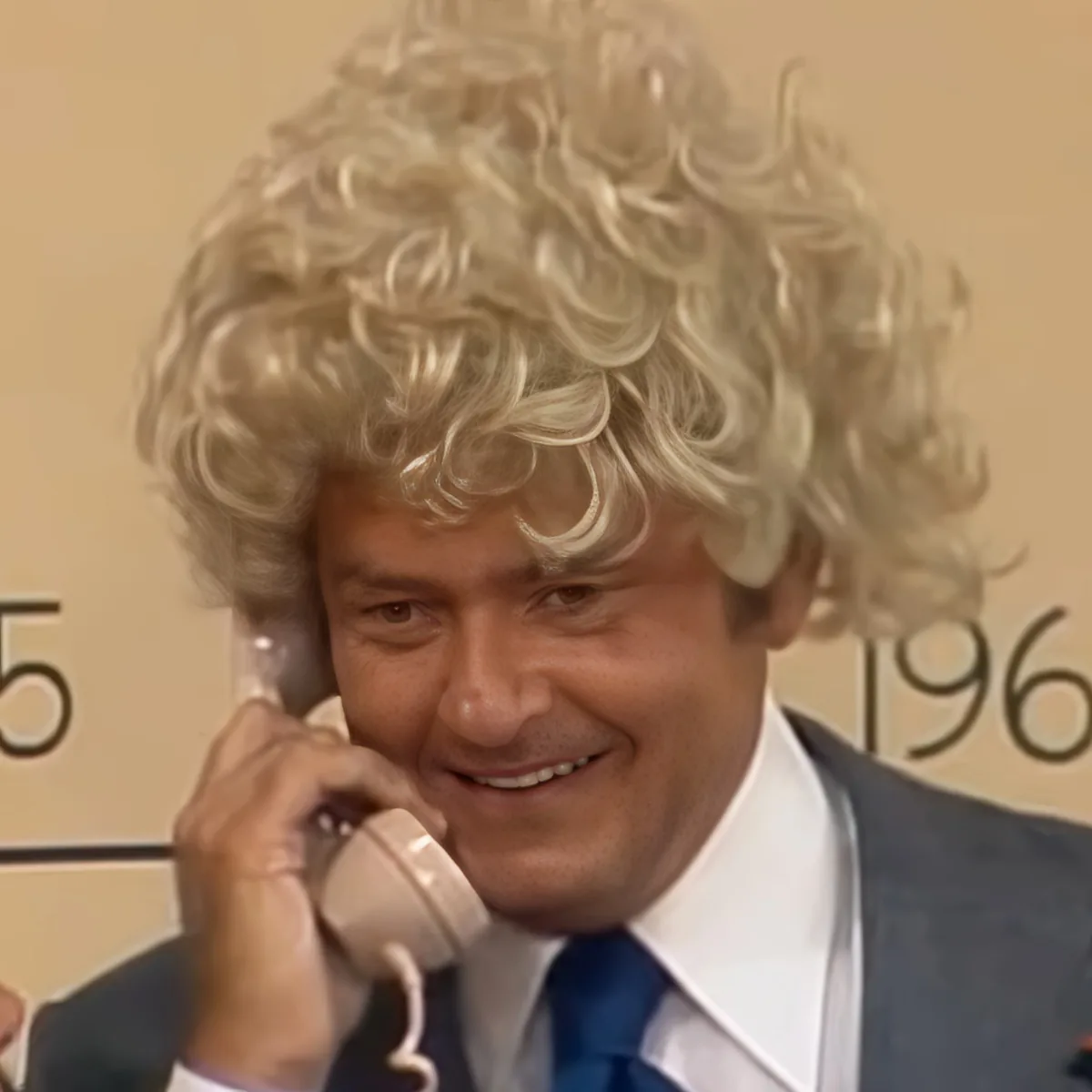 A man with curly blonde hair, wearing a suit and tie, is holding a phone to his ear and smiling.