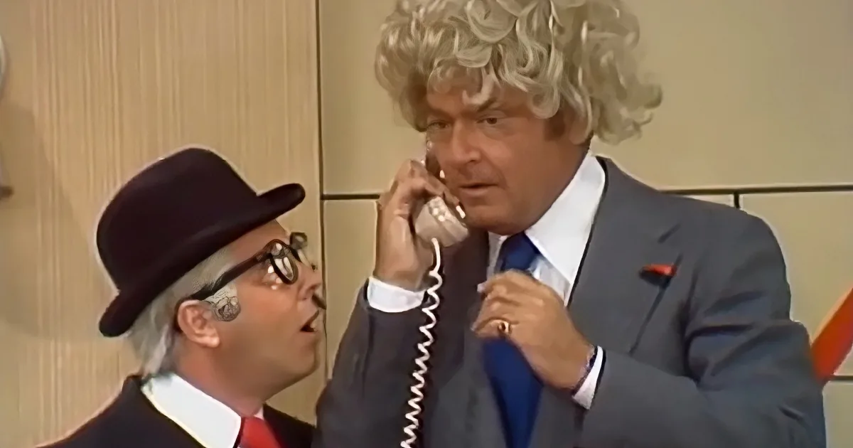 A man in a suit and blonde wig is talking on the phone while another man in a top hat looks on.