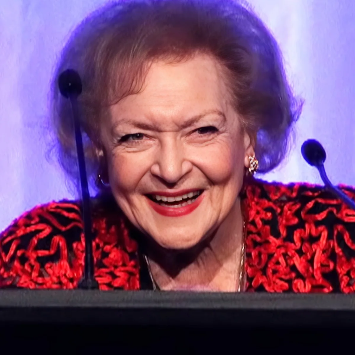 An older woman with short hair, wearing a red patterned jacket, smiles while speaking into a microphone.