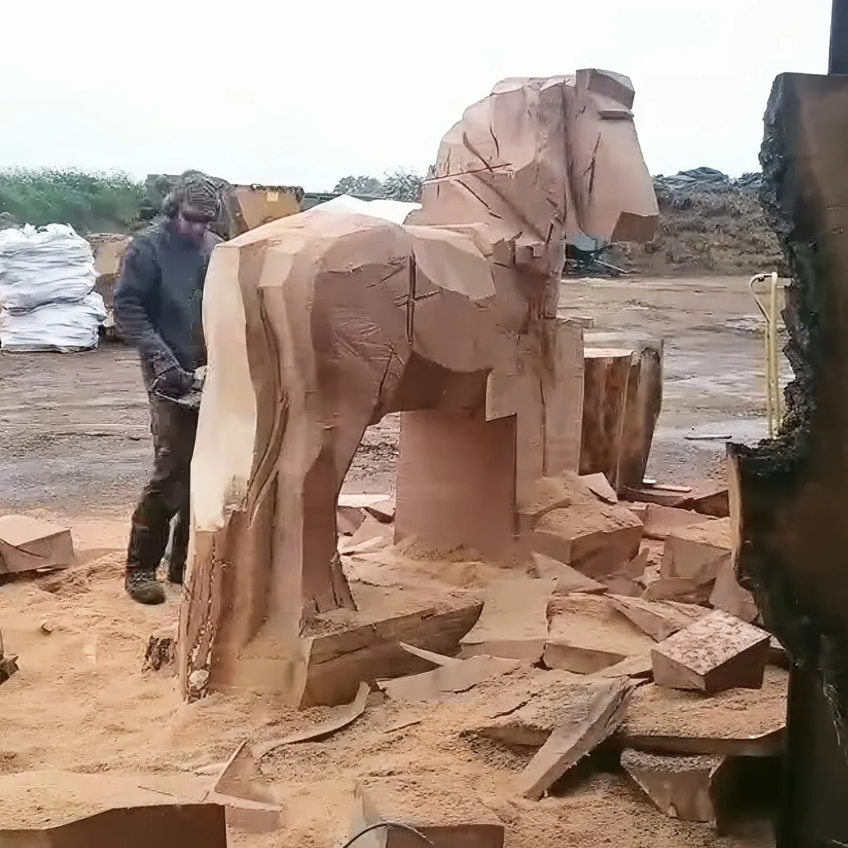 A sculptor works on a large wooden elephant sculpture, surrounded by wood shavings and tools.