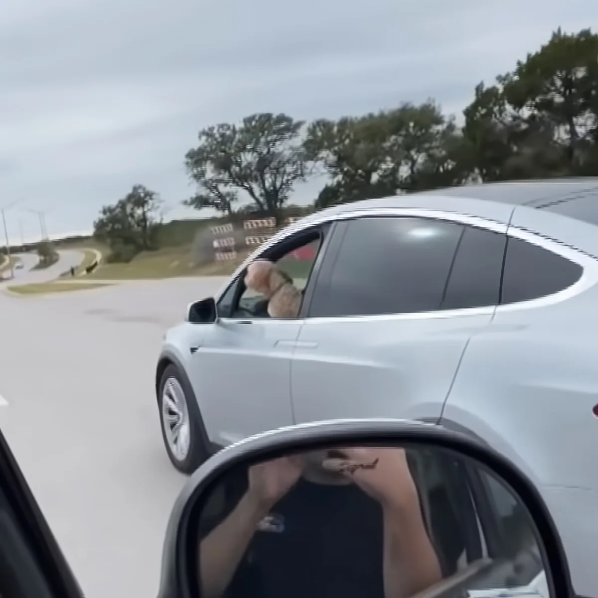 A white Tesla Model Y is driving on a road with a person in the side mirror holding a phone.