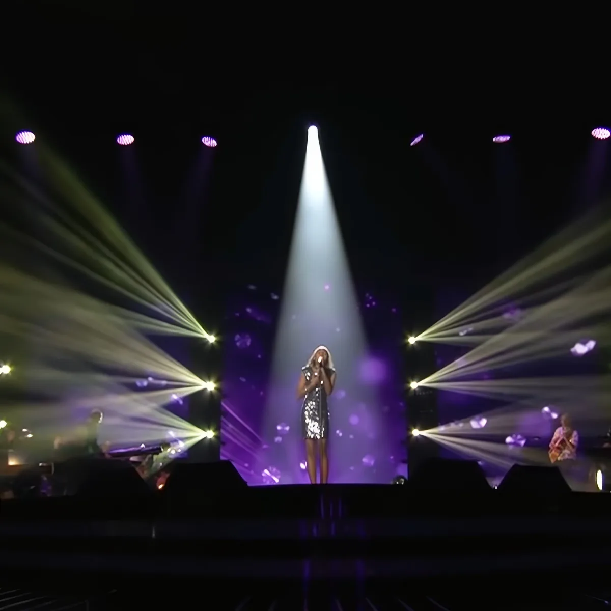 A singer in a sequined dress stands under a spotlight, surrounded by colorful stage lights.