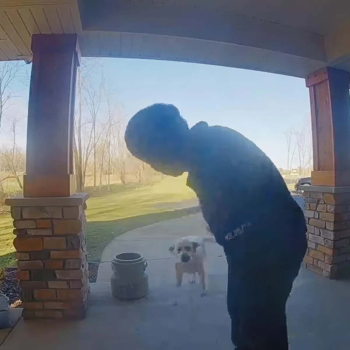 A person bends down on a porch, interacting with a small dog near a trash can.