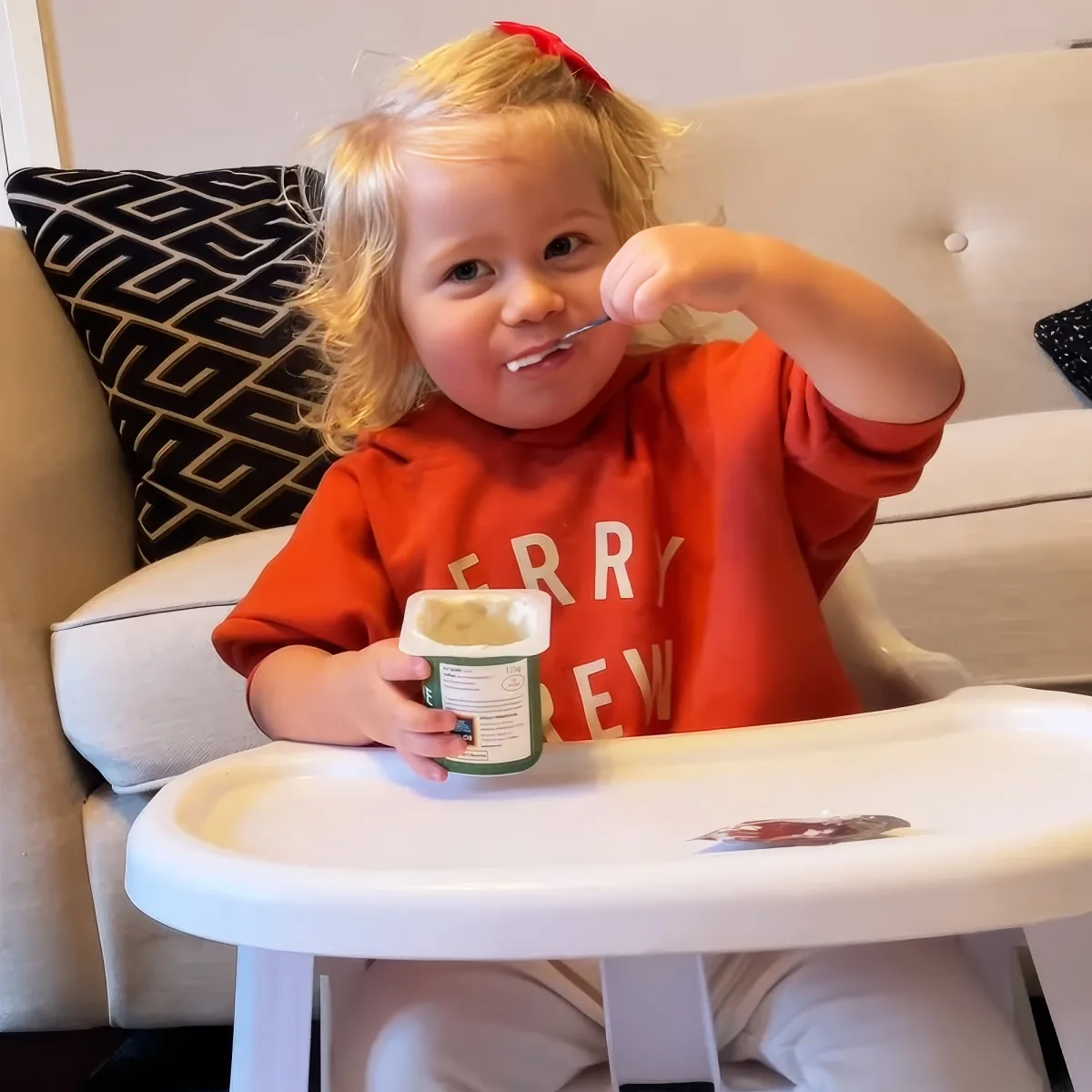 A toddler in a red shirt sits in a high chair, holding a yogurt container and eating with a spoon.