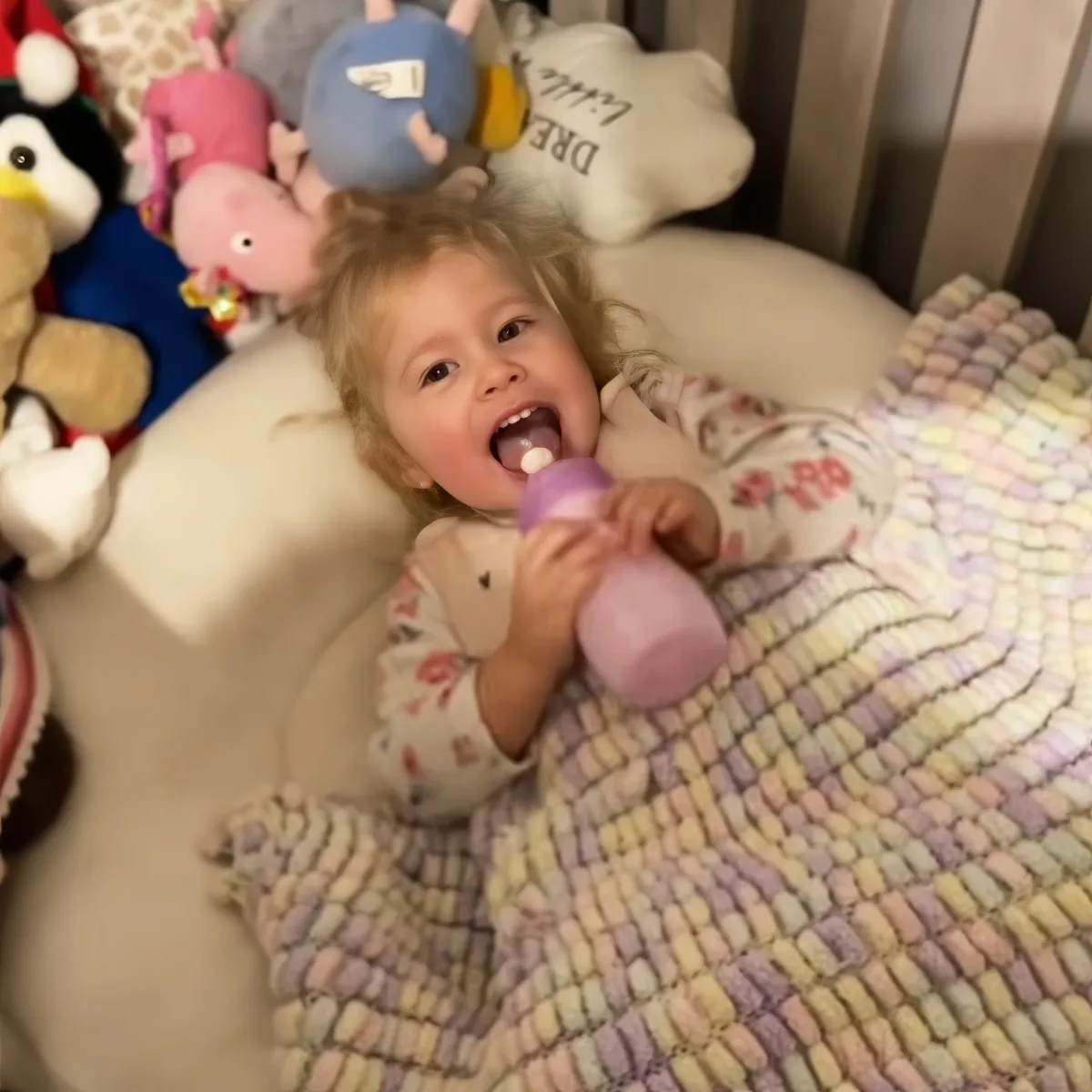 A happy toddler lies in a crib with a pastel blanket, holding a pink sippy cup and surrounded by stuffed animals.