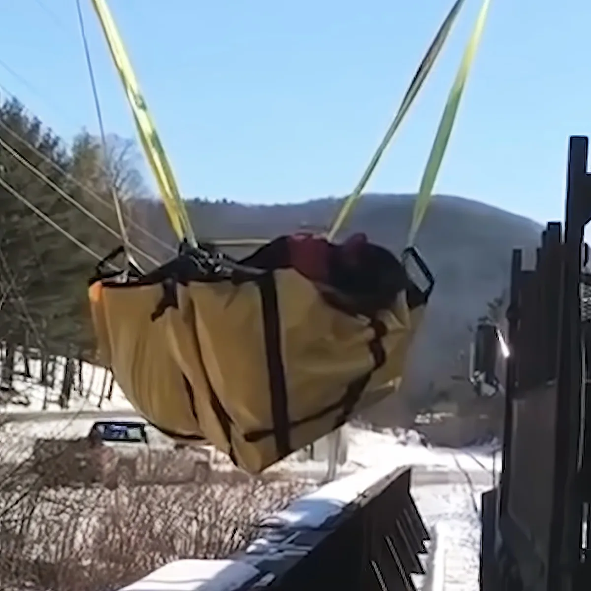 A person is suspended in a hammock-like seat, secured by yellow straps, with a snowy landscape and trees in the background.