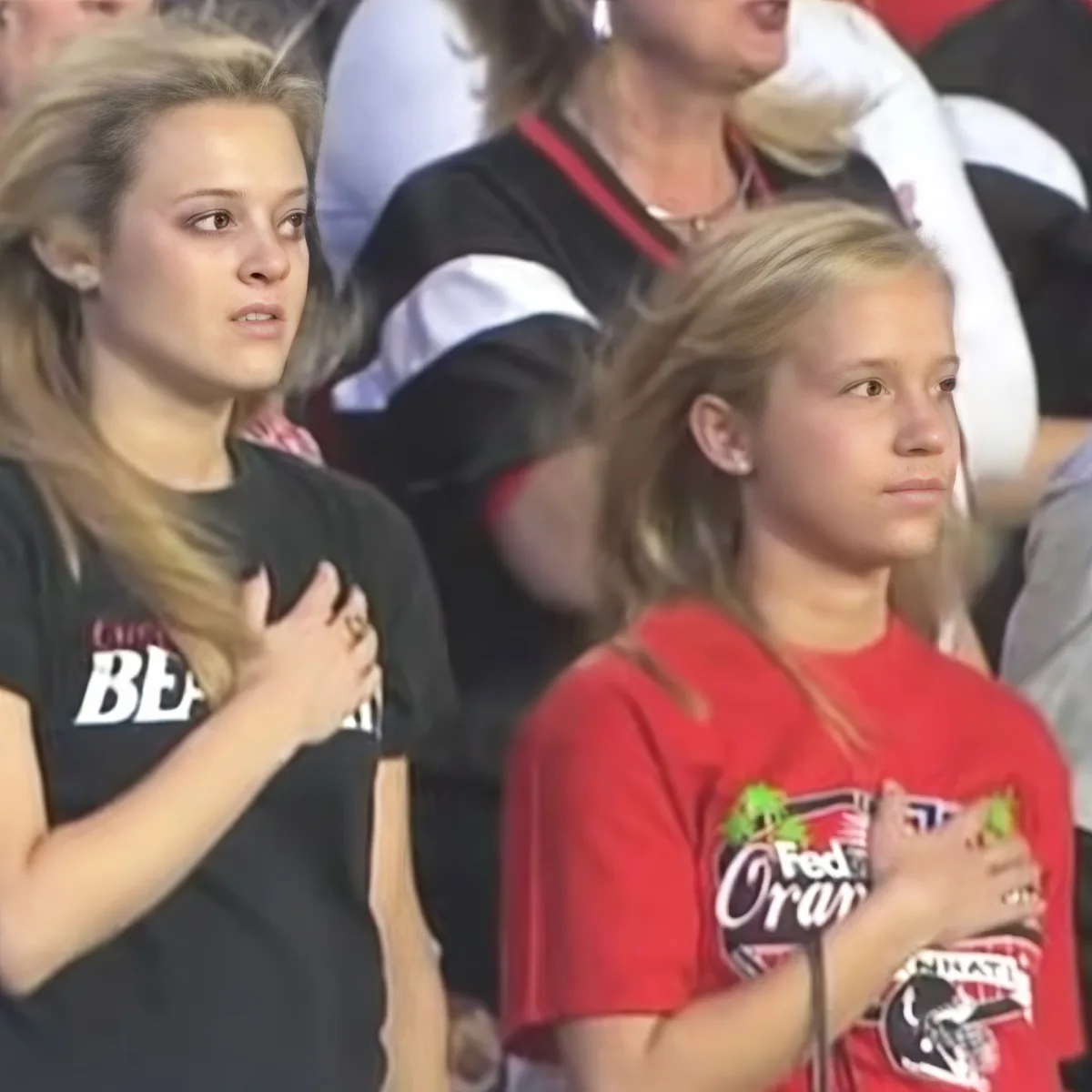 A young girl in a red shirt holds her hand over her heart, standing next to another person in a black shirt.
