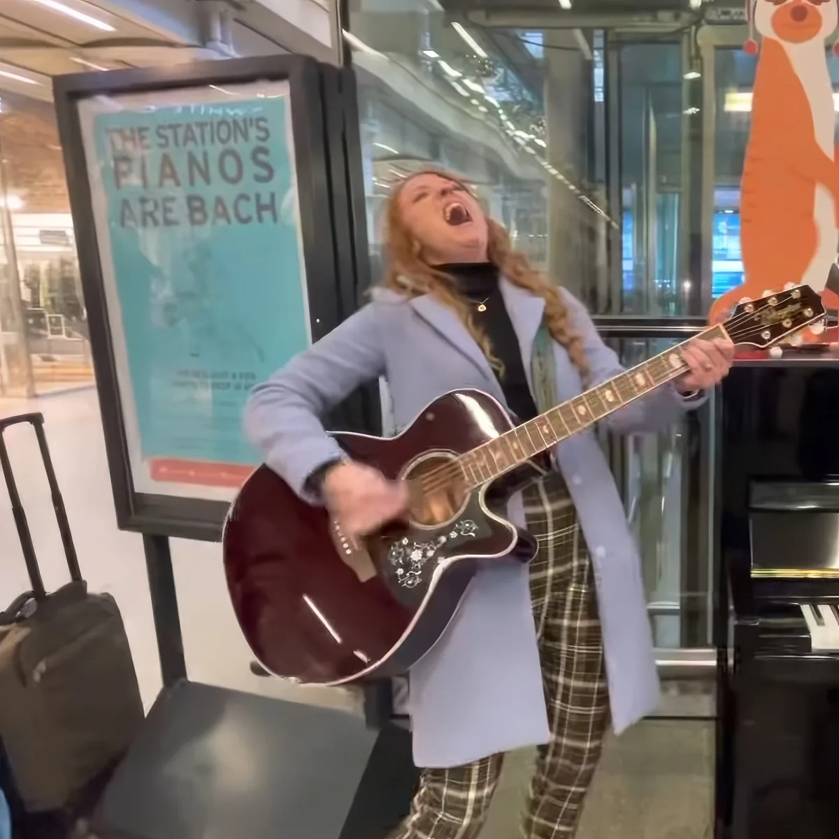 A person in plaid pants and a light blue coat plays an acoustic guitar in front of a sign that says "The Station's Pianos...