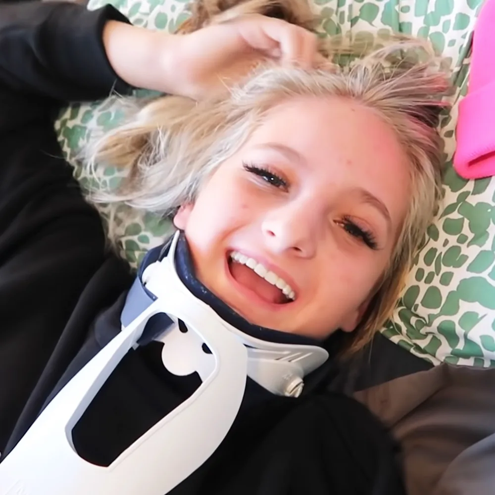 A person with blonde hair and a white neck brace is lying down, smiling, with a green patterned pillow behind them.