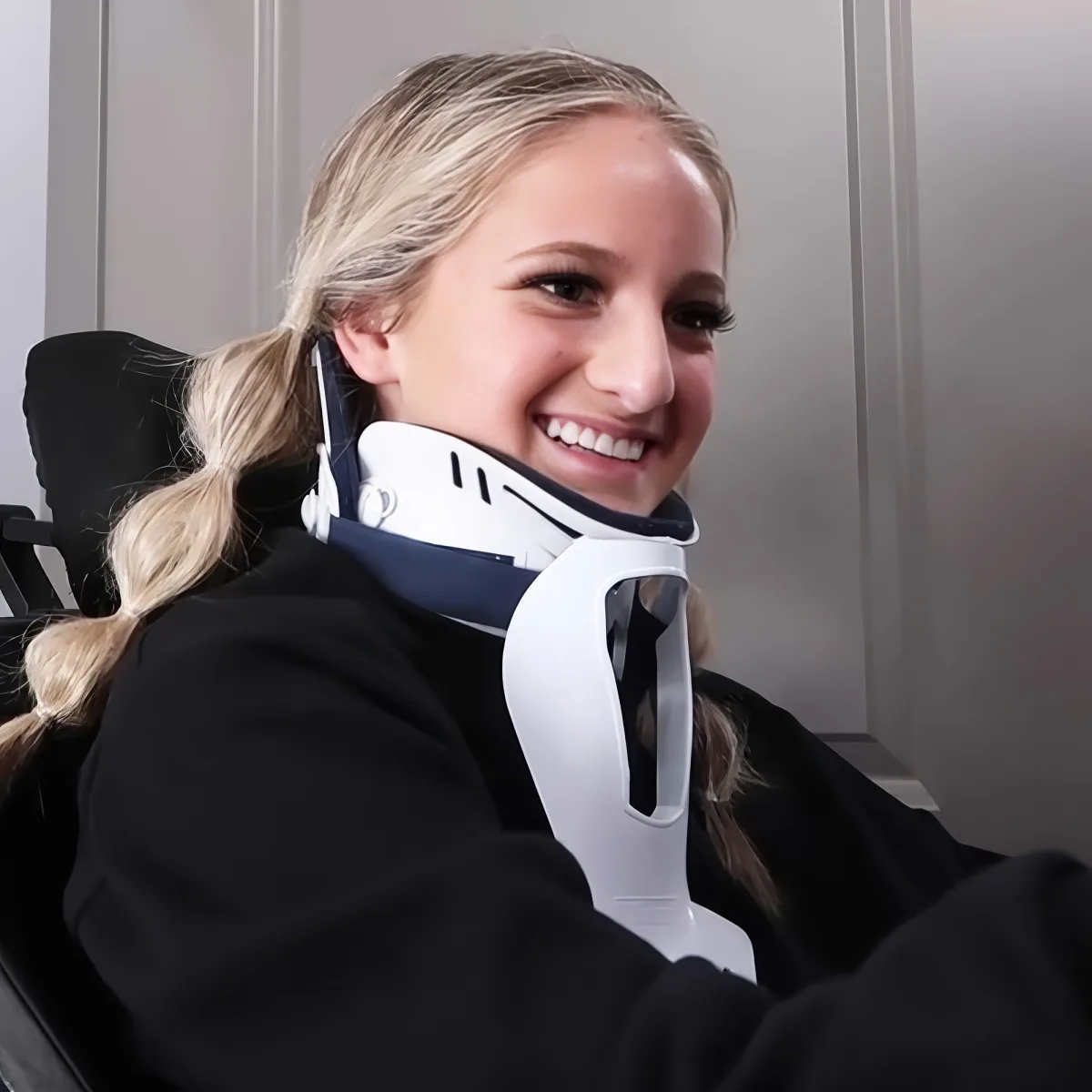 A person with long blonde hair smiles while wearing a white and blue neck brace, seated in a black chair.