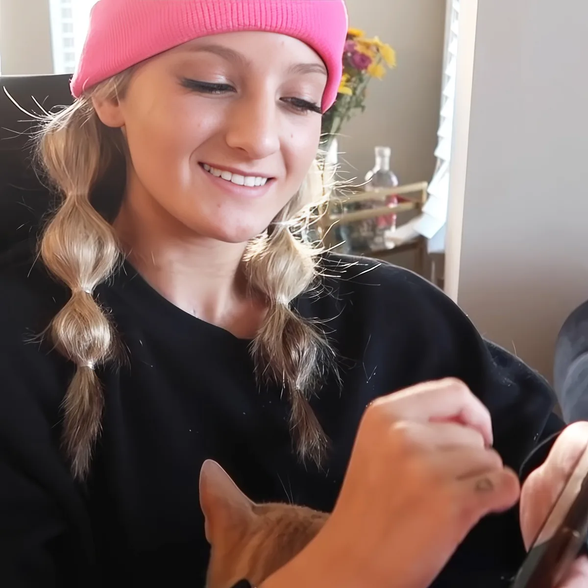 A smiling person wearing a pink beanie and black shirt, holding a phone, with a cat partially visible.