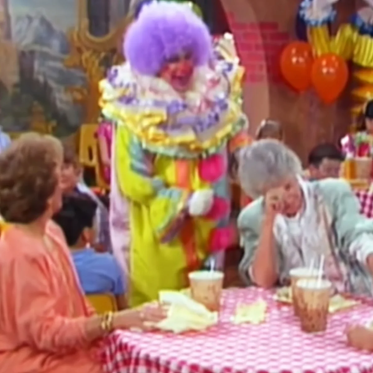 A clown in a colorful outfit stands at a table with a red-and-white checkered tablecloth, surrounded by children.