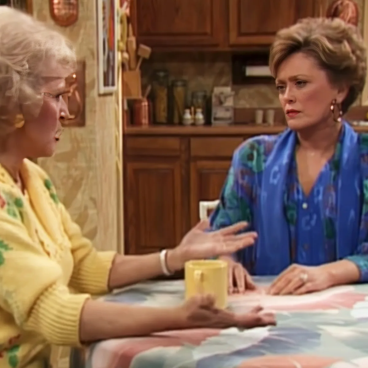 Two women in a kitchen setting, one gesturing while holding a yellow mug, the other listening attentively.