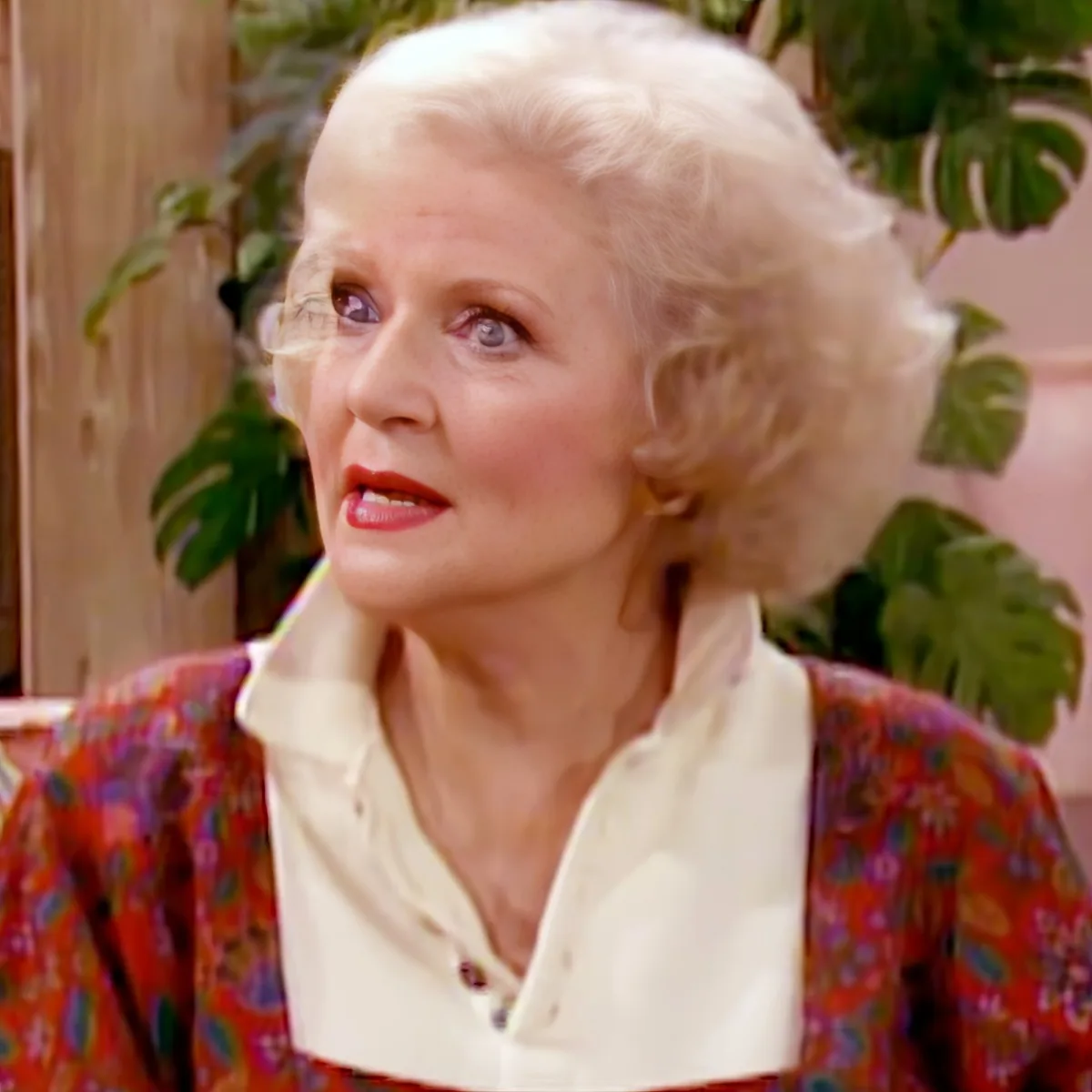 An older woman with white hair, wearing a floral jacket and white shirt, is looking to the side.