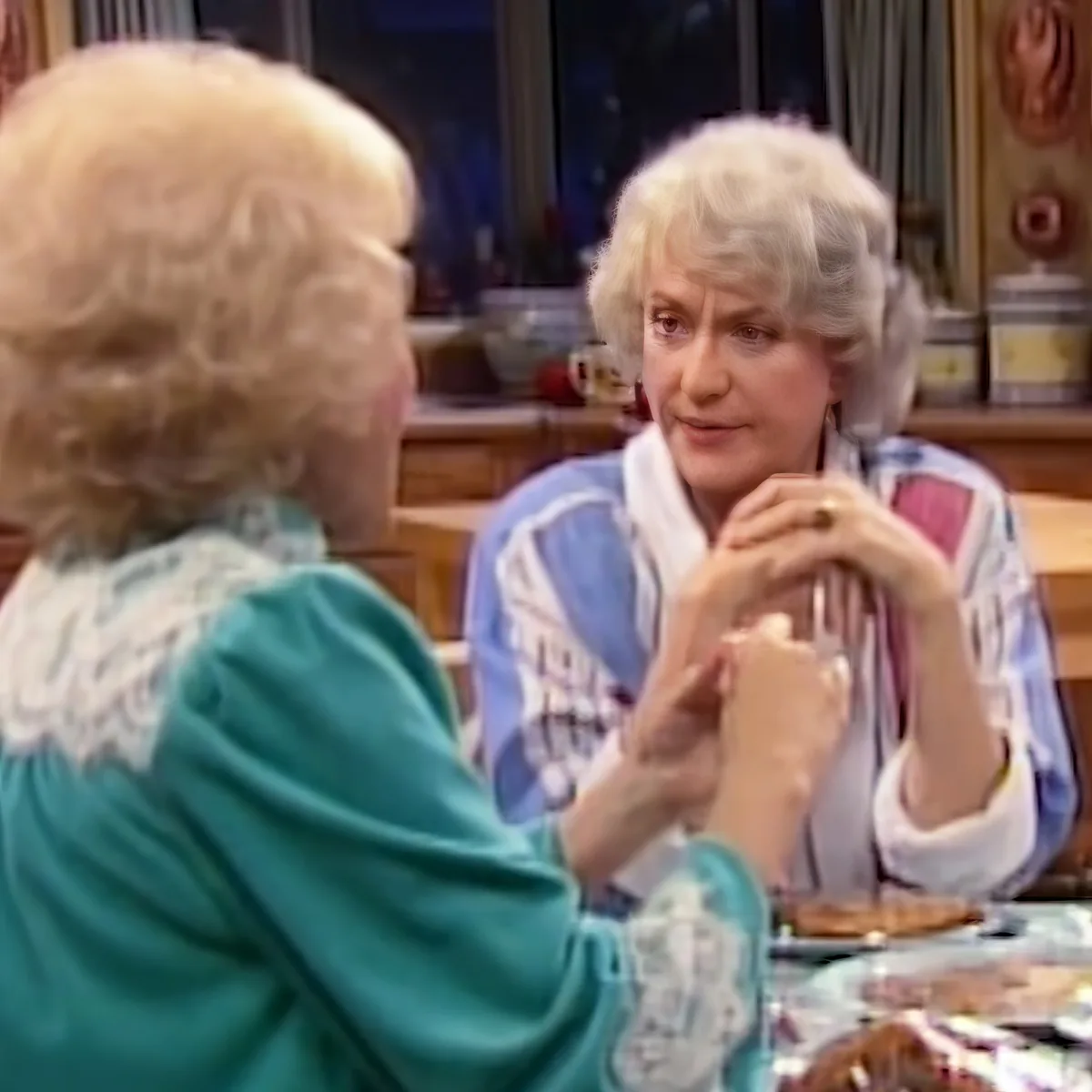 Two women are seated at a table, one in a teal sweater holding a glass, the other in a patterned shirt with a white...