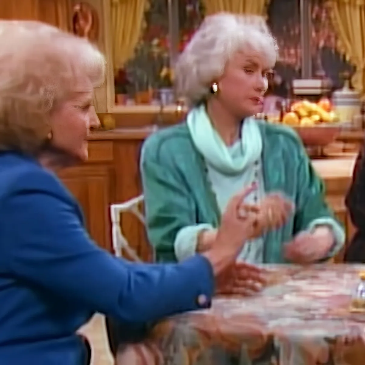 Two women are seated at a table, one in a blue jacket gesturing, in a cozy kitchen setting.