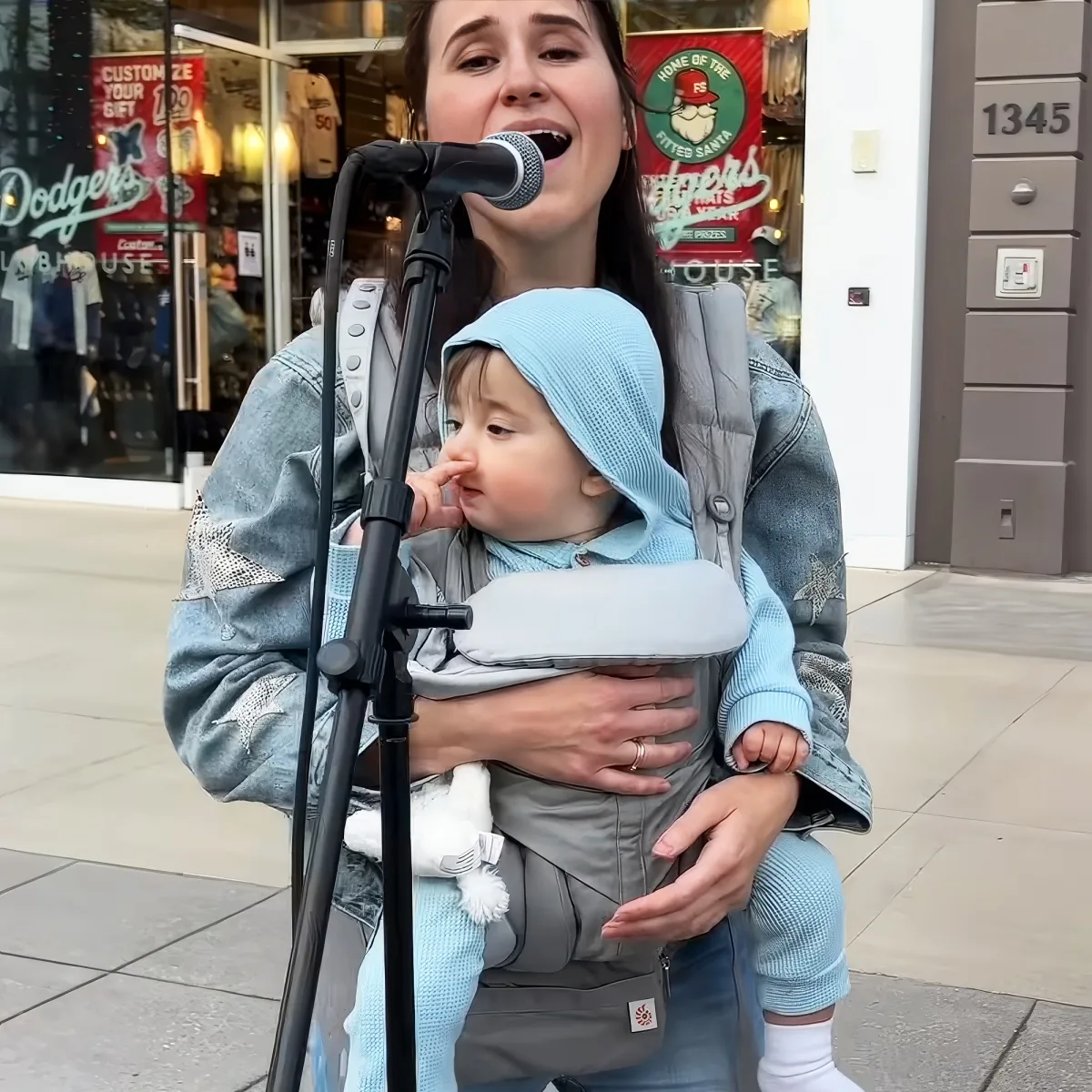 A woman sings into a microphone while carrying a baby in a carrier, with a storefront in the background.